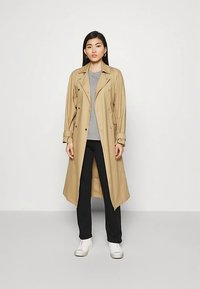 Beiger Trenchcoat mit doppelreihigem Design, Knopfdetails und verstellbaren Manschetten, getragen über einem grauen Oberteil und schwarzen Hosen. Weiße Sneakers.