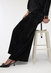 Pantaloni a palazzo in velluto nero con una consistenza morbida, abbinati a décolleté in vernice nera con tacco a slingback e dettagli decorativi a fiocco.