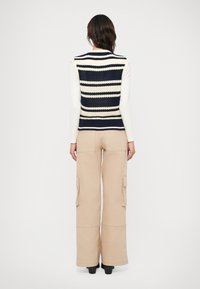 Colete de malha com riscas horizontais em azul-marinho e creme, decote redondo, usado sobre uma blusa branca de mangas longas. Calças cargo largas em bege.