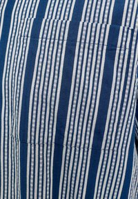 Kék-fehér, függőlegesen csíkozott anyag, varrott négyzet alakú zsebbel, amelyen egyező csíkminta és varrott szegélyek találhatók.