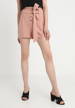 Pantaloni corti in lino rosa chiaro con vita alta, bottoni frontali e cintura legata. Abbinati a sandali neri con tacco a blocco e strap.