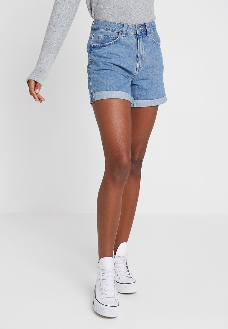 Jeansshorts med en ljusblå tvätt, uppvikta benslut och framfickor. Bärs med vita högskurna sneakers mot en neutral bakgrund.