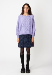 Pull en maille de lilas avec un motif géométrique, associé à une jupe à motif bleu foncé. Le mannequin porte des bottes noires en maille jusqu'au genou.