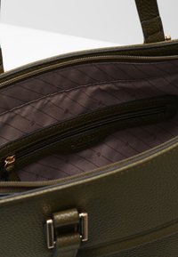 Sac en cuir grainé vert olive avec une fermeture éclair, doté d’un intérieur doublé en marron et d’une poche décorative zippée. Orné de ferrures dorées.