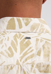Camisa de manga corta con una tela blanca que presenta grandes patrones de hojas abstractas en verde claro; incluye una pequeña etiqueta metálica de la marca cerca del cuello.