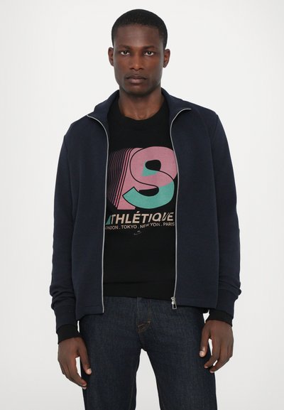Giacca zip-up blu navy sopra una t-shirt nera con grafica che presenta testo multicolore. Jeans in denim scuro con cuciture arancioni. Tessuto liscio, design aderente.
