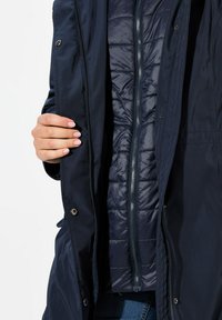 Giacca esterna blu navy con una texture liscia e opaca e chiusure a bottone, che rivela un rivestimento interno trapuntato con una finitura lucida.