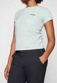 T-shirt en coton vert clair à manches courtes, col rond, avec un logo "Patagonia" noir sur la poitrine gauche. Porté avec un pantalon sombre.