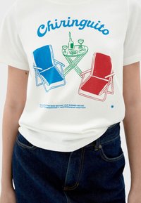 White cotton t-shirt featuring blue and red illustrated beach chairs, a green table, drinks, and the text "Chiringuito" in bold typography.