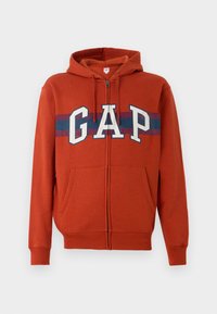 Crvena zip-up dukserica zahtjeva od pamuka s flis teksturom. Ima bijeli "GAP" logo okružen plavim i bordo prugastim pojasom. Dva prednja džepa.