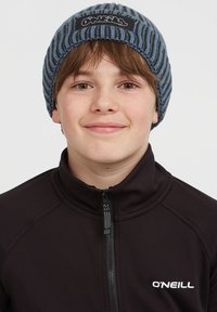Bonnet en tricot à rayures bleues et grises avec un patch logo noir. Porté par une personne dans un haut zippé noir. Matériau doux et texturé.