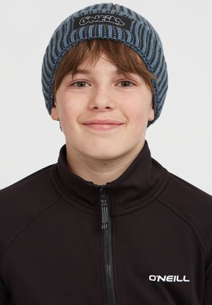 Gorro de punto en rayas azul y gris con un parche de logo negro. Lo lleva puesto una persona con una sudadera negra con cremallera. Material suave y texturizado.
