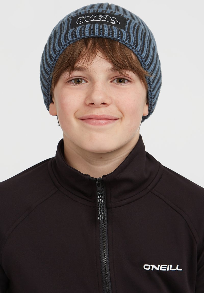 Bonnet en tricot à rayures bleues et grises avec un patch logo noir. Porté par une personne dans un haut zippé noir. Matériau doux et texturé.
