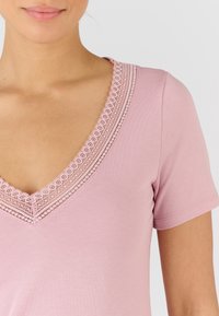 Camicia rosa a maniche corte con scollatura a V e dettagli in pizzo elaborato lungo il collo. Realizzata in tessuto morbido e liscio.