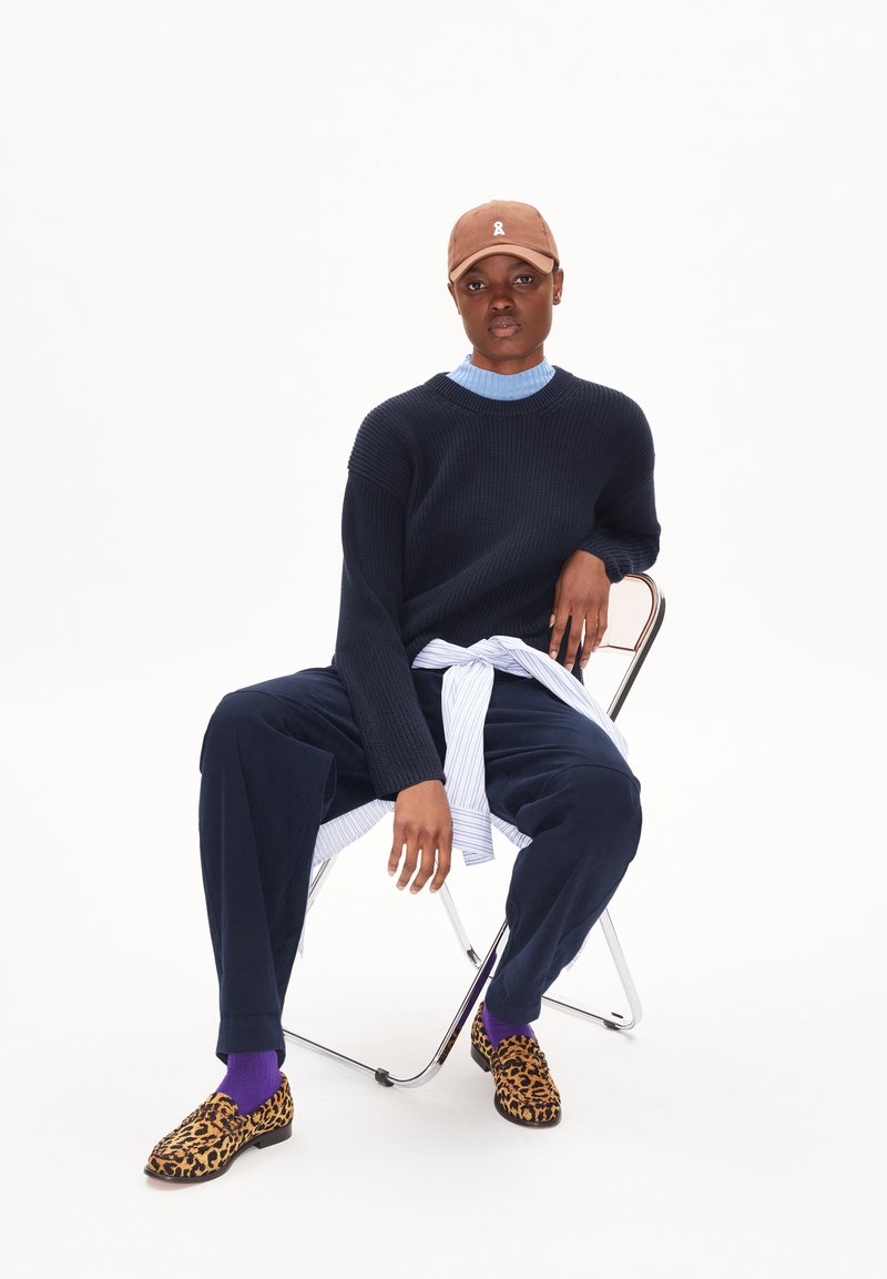 Marineblauer gerippter Pullover über einem hellblauen Hemd, kombiniert mit marineblauen Hosen, lila Socken und Schuhen im Leopardenmuster. Das Model sitzt auf einem Stuhl.