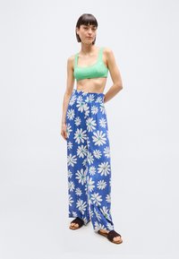 Νεαρή γυναίκα στέκεται φορώντας πράσινο κοντό τοπ, μπλε παντελόνες με λευκά σχέδια γερανιού και σκούρα πλεκτά σανδάλια χωρίς λουράκια, σε απλό φόντο.