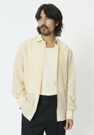 Homme aux cheveux longs jusqu'aux épaules et à la barbe, portant une chemise à rayures jaunes et blanches par-dessus un débardeur blanc et un pantalon noir, regardant vers la gauche.