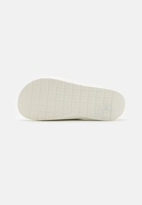 Semelle de chaussure blanche plate avec motif en quadrillage et petit détail circulaire près du talon sur un fond clair uni.