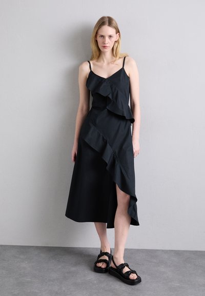 Rochie neagră cu volane suprapuse, bretele subțiri și o fante laterală. Asortată cu sandale negre masive cu un design legat.