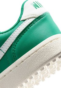 Zielone skórzane trampki z białymi akcentami, charakteryzujące się teksturowaną gumową podeszwą i przeszyciami. Zaokrąglony czubek i niska sylwetka.