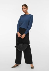 Femme portant un haut à manches longues bleu, pantalon large noir, escarpins imprimé zèbre, tenant un sac à main structuré noir, posant devant un fond blanc.
