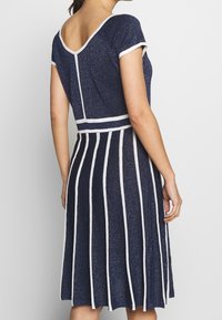 Marinblå klänning med korta ärmar, rygg med V-form, vita randiga detaljer, figursydd midja och en utsvängd kjol som visar ett strukturerat, glittrande mönster.