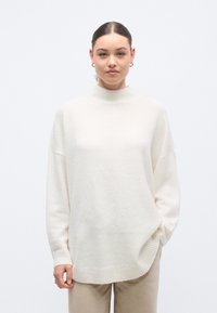 Kremo spalvos didelio dydžio megztinis su aukštu kaklu, nuleistais pečiais ir akordeono rankogaliais. Minkšta tekstūra ir laisvas kirpimas. Derinamas su šviesiai smėlio spalvos kelnėmis.