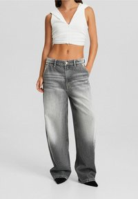 Nainen, joka pitää päällään valkoista ilmanhihoista, lyhyttä vetoketjullista toppia ja korkeavyötäröisiä haalistuneita harmaita leveälahkeisia farkkuja, seisoo kädet taskuissa.