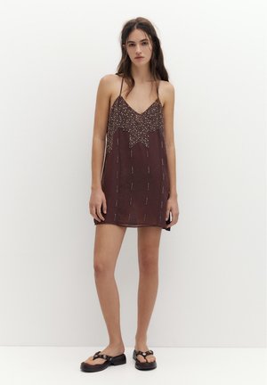 Fiatal nő áll, barna, ujjatlan mini ruhát visel csillagmintával és fekete platform szandállal, fehér fal előtt.