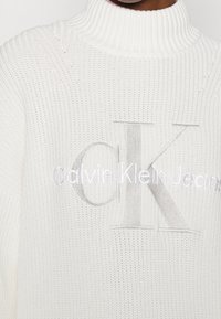Pull en tricot blanc avec un col montant, présentant des lettres grises en relief "CK" et "Calvin Klein Jeans" sur le devant. Design de tissu texturé.