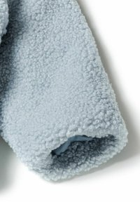 Ljust blått fleece-tyg, strukturerat med en plyschig, fluffig yta; den uppvikta ärmslutet visar en solid inre foder.