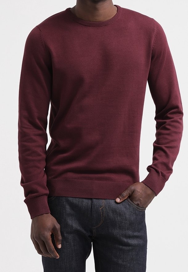 Personne portant un pull bordeaux uni à manches longues et un jean bleu foncé, avec une main dans la poche et un arrière-plan neutre.