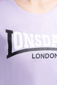 Detailný záber na levanduľovú košeľu s veľkým bielym a čiernym nápisom "Lonsdale" a menším čiernym nápisom "London" pod ním.