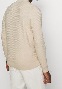 Mann, der einen hellbeigen, strukturierten Strickpullover und off-white Hosen trägt, von hinten vor einem einfarbigen, hellen Hintergrund gezeigt.