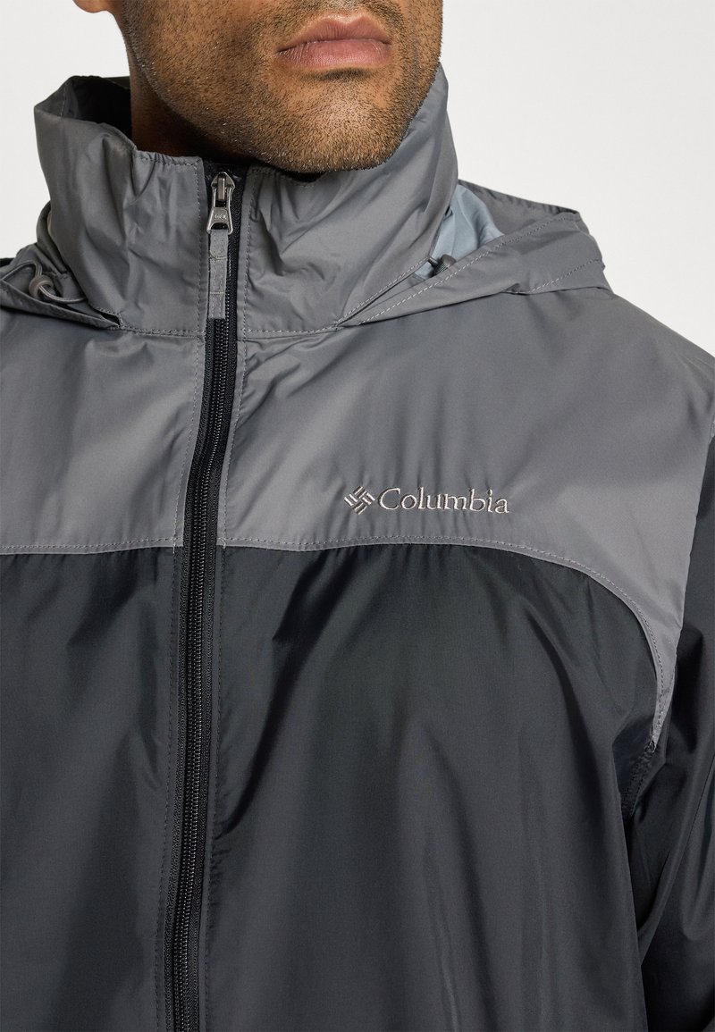 Chaqueta gris y negra con cierre delantero y cuello alto. Presenta un logotipo en el pecho y una capucha. Fabricada con material liso y resistente al agua.