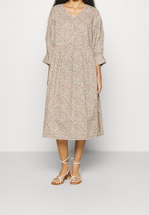 Blumenkleid in Beige mit pinken und grünen Mustern. Hat einen V-Ausschnitt, eine geraffte Taille und Dreiviertelärmel. Kombiniert mit Riemchensandalen.