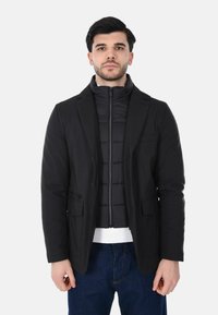 Giacca nera a strati con stile imbottito, chiusura frontale con zip, tasche e design strutturato da blazer, indossata sopra un maglione bianco.