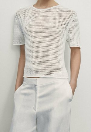 Personne portant un haut tricoté blanc à manches courtes avec un motif texturé et un pantalon blanc taille haute, mains dans les poches.