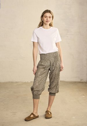 Nő áll bent, fehér rövid ujjú inget, mintás, laza, rövidített nadrágot és barna bebújós cipőt visel.