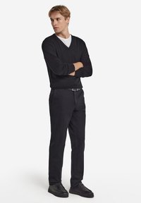 Svart tröja med V-ringning, lagd över en vit T-shirt. Svarta slim-fit byxor och mörka sneakers. Slät tyg med en figurnära design.