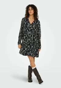 Robe à motifs noir et blanc avec un décolleté en V, des manches longues et un ourlet évasé, associée à des bottes brunes jusqu'aux genoux et une chaussette légère.
