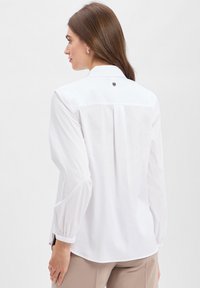 Vrouw met lang bruin haar, gekleed in een witte blouse met lange mouwen en beige broek, met haar rug naar de camera gericht tegen een effen witte achtergrond.