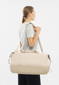 Beige Duffelbag mit glatter Textur, ausgestattet mit zwei oberen Griffen und einem abnehmbaren Schultergurt. Dezentes Logo auf der Vorderseite.