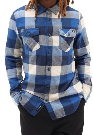 Personne portant une chemise à carreaux bleue, blanche et grise à manches longues avec deux poches poitrine boutonnées, mains dans les poches d'un pantalon noir.