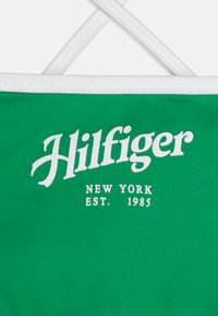 Tessuto verde con accenti bianchi che presenta il testo "Hilfiger" e "NEW YORK EST. 1985" in caratteri in grassetto. Texture liscia, design minimalista.