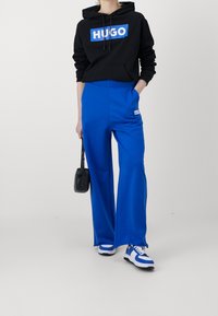 Sudadera negra con el logotipo "HUGO" en blanco, combinada con pantalones de chándal anchos azules con un pequeño parche de logo, y zapatillas azul y blancas.