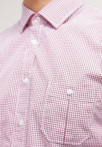 Camisa de botones con estampado de cuadros rojos y blancos, con cuello y bolsillo en el pecho, botones blancos y material de algodón texturizado.