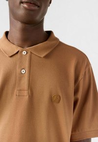 Polo marrone realizzata in tessuto strutturato, con una patta a tre bottoni e un piccolo logo in rilievo sul petto.