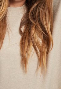 Maglione beige chiaro con un girocollo alto, texture morbida e vestibilità leggermente ampia, caratterizzato da capelli lunghi e ondulati in tonalità bionde.