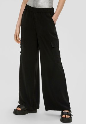 Zwarte wijde broek gemaakt van een zachte stof, met zijzakken en een elastische tailleband. Draagt bij chunky zwarte sandalen.